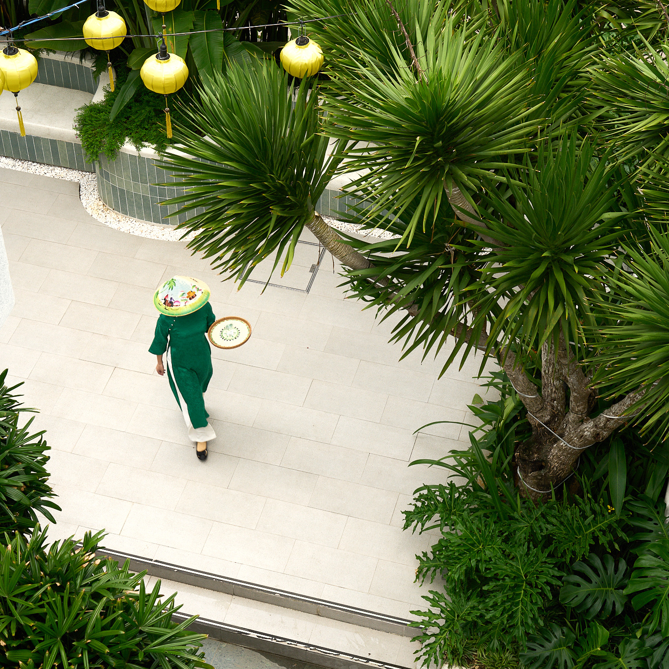 Staff member in a green áo dài walking beneath yellow lanterns and palms