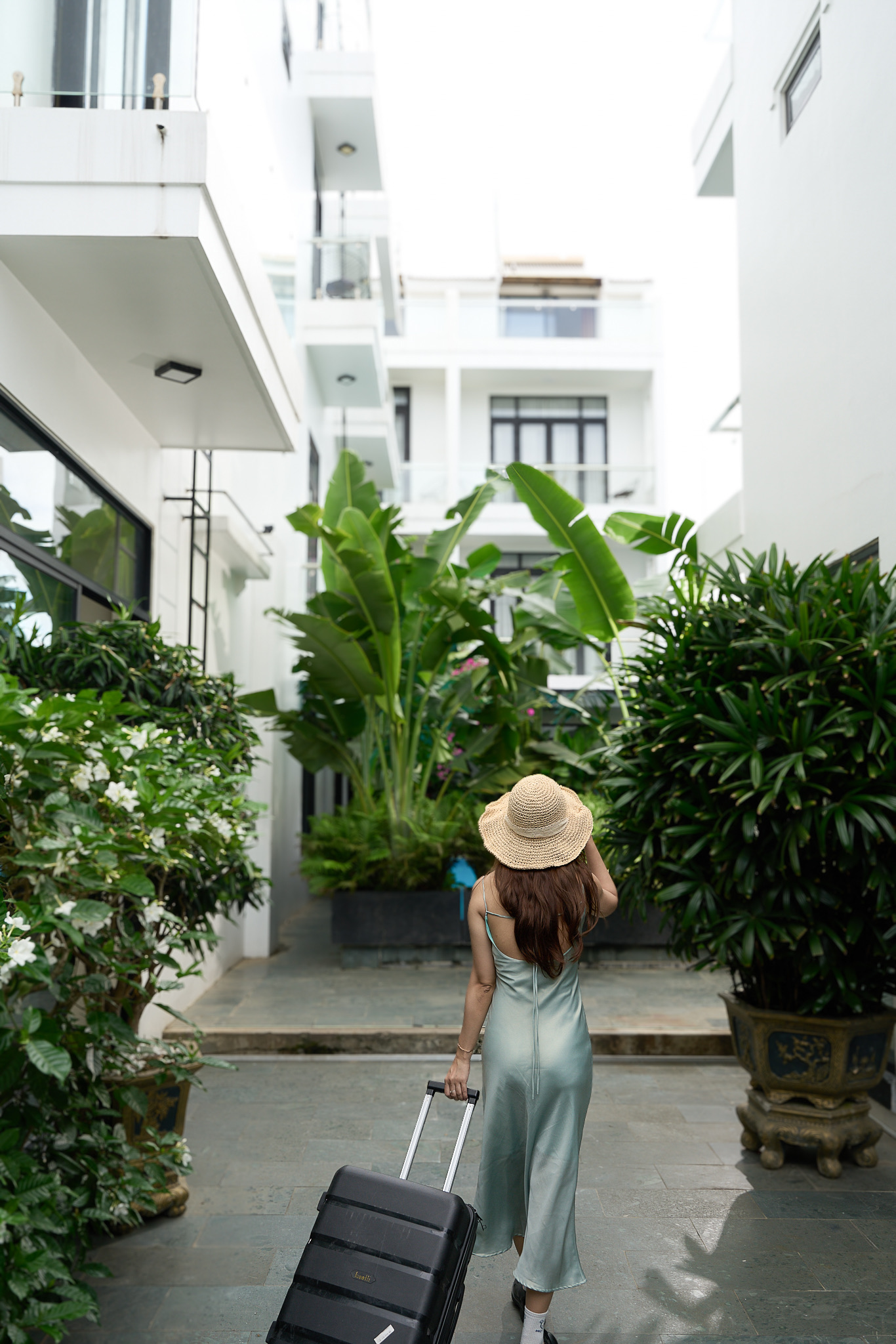 A guest in a sun hat wheeling a suitcase past banana palms toward the villa
